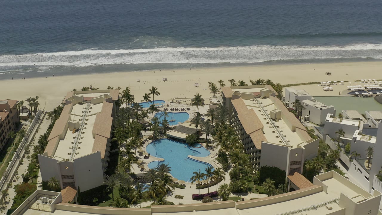 High aerial view in Los Cabos, Mexico, looking over tan-roofed resort buildings, curved blue pools, palm trees, beach sand, surf, and ocean with no visible people.