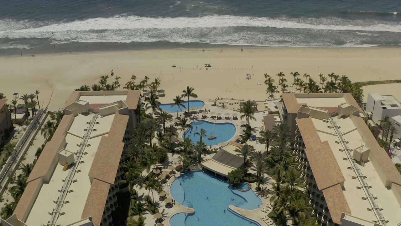 Overhead view of a Los Cabos beach resort with blue pools and palm trees in the foreground, broad sand beyond, white surf at the top, and no discernible people.