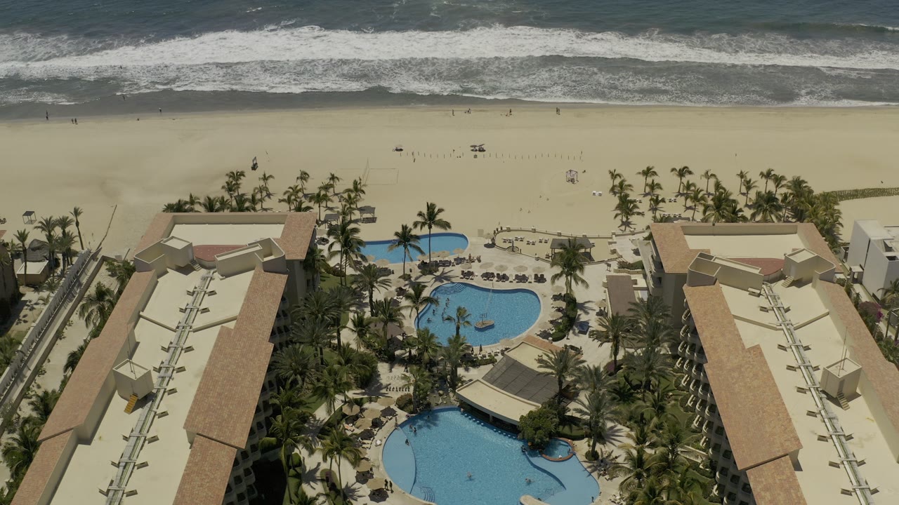 Bird's-eye view of resort pools, palms, open beach, and surf.