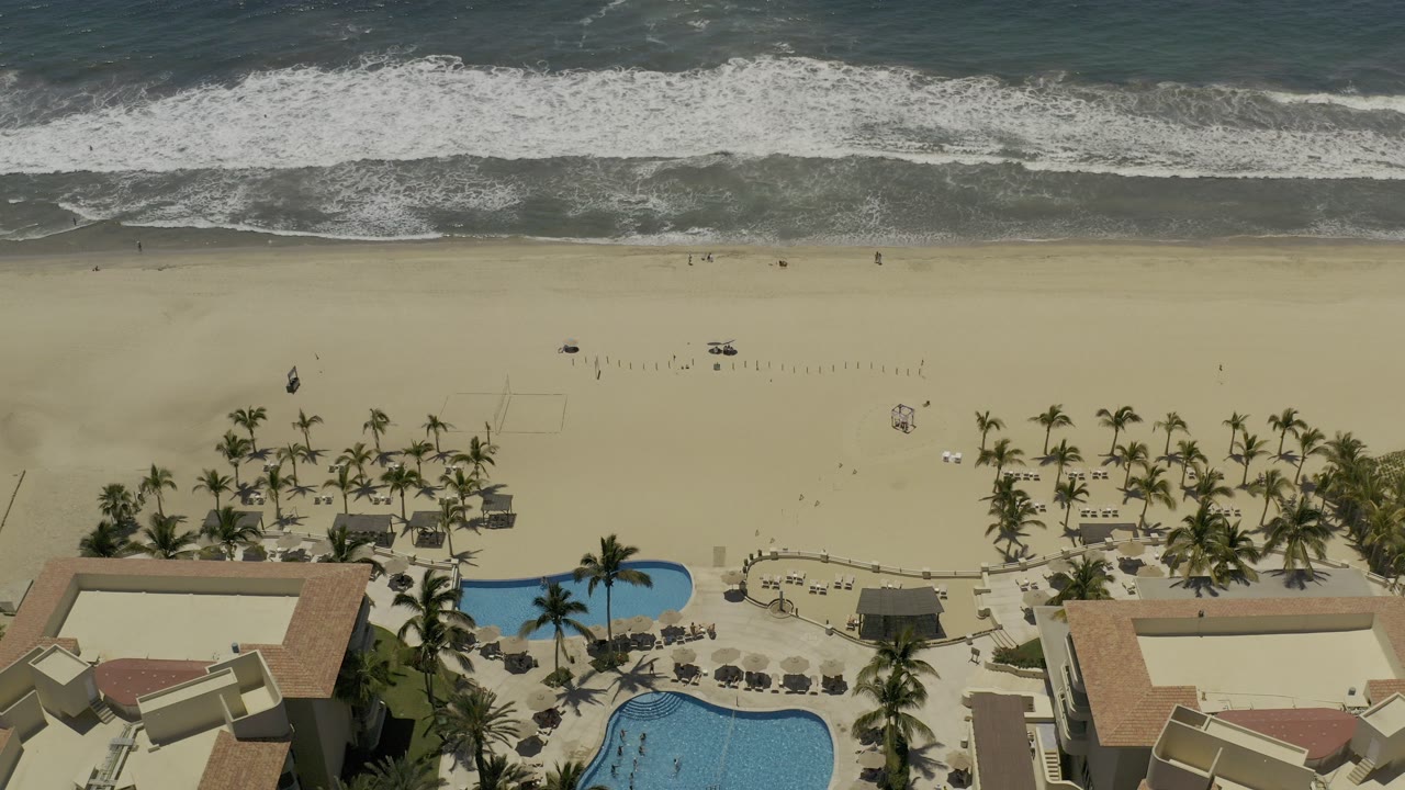 Aerial frame over a Los Cabos beach shows white surf above wide tan sand, palm clusters, blue pool water, and resort buildings along the lower edge with no clearly visible people.