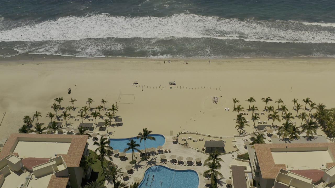 A second Los Cabos aerial frame shows waves reaching the upper shoreline, a broad empty-looking sand band, palms, loungers, and curved blue pool shapes near the bottom with no clearly visible people.