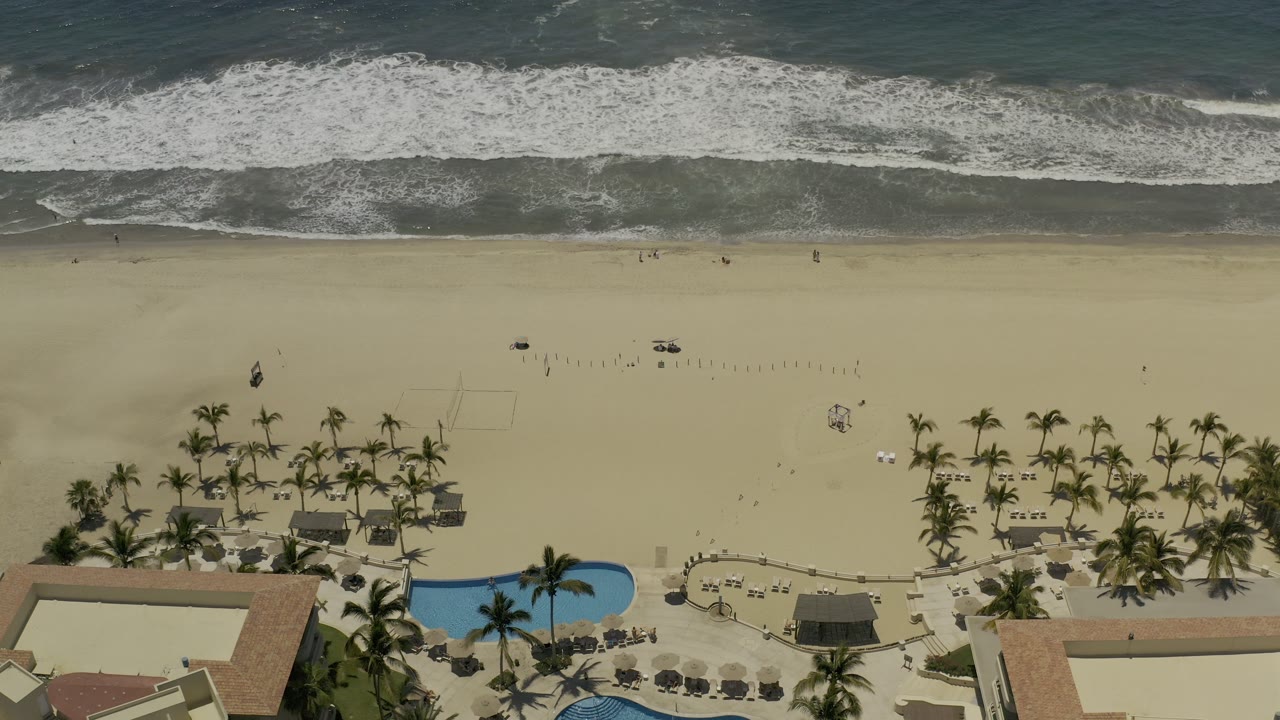 Drone-like view of shoreline, beach palms, resort pools, and buildings.
