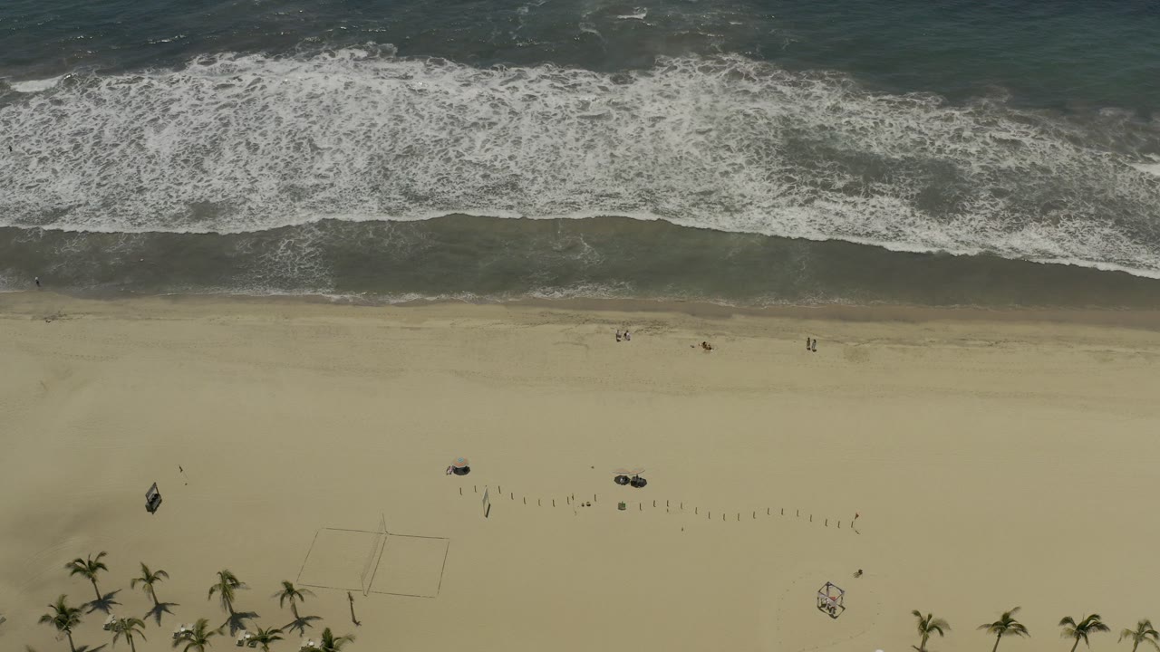 Overhead view of a Los Cabos beach at 0:00, with white surf breaking across dark blue-green water above pale sand and no visible people.