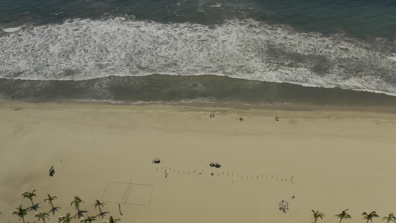 Aerial view of the Los Cabos shoreline at 0:01, showing a broad white wave band, smooth sand below, faint rectangular beach markings, and no visible people.