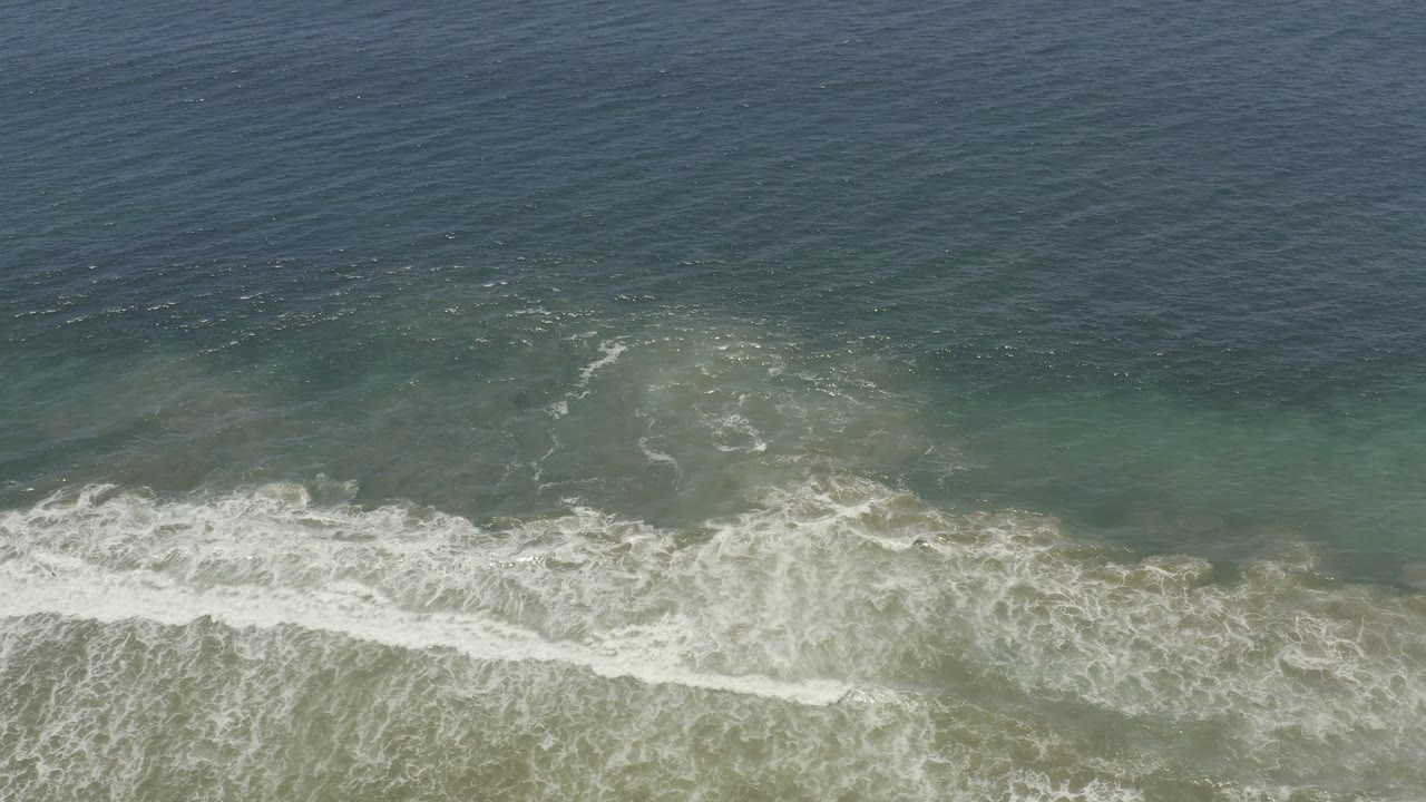 Aerial ocean frame from Los Cabos, Mexico, showing a broad white wave line rolling through green-blue water with no visible people.