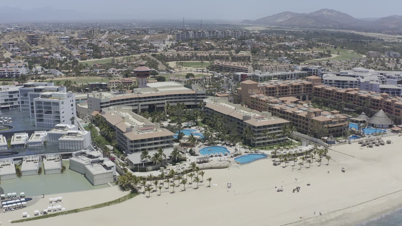 Opening aerial frame of a large Los Cabos resort complex beside pale beach and shallow water, with pools, white and tan buildings, distant hills, hazy sky, and no visible people.