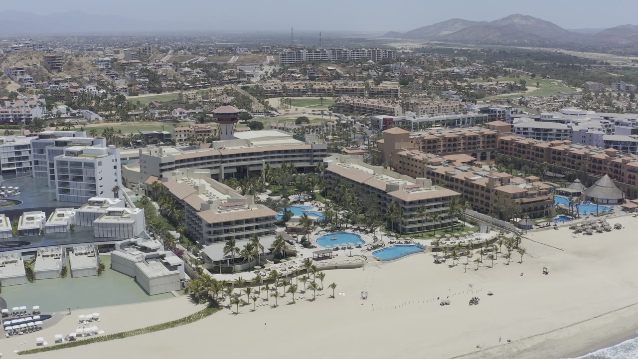 Wide aerial view of beachfront buildings, sand, surf, pools, and distant hills.