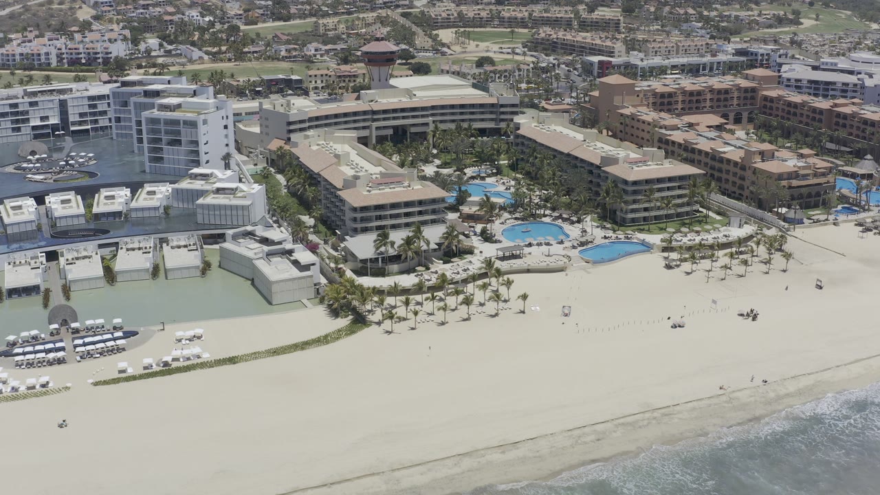 Aerial frame over Los Cabos, Mexico showing a broad sandy beach, a strip of surf at the lower edge, palm-lined pool areas, white and tan buildings, and no visible people.