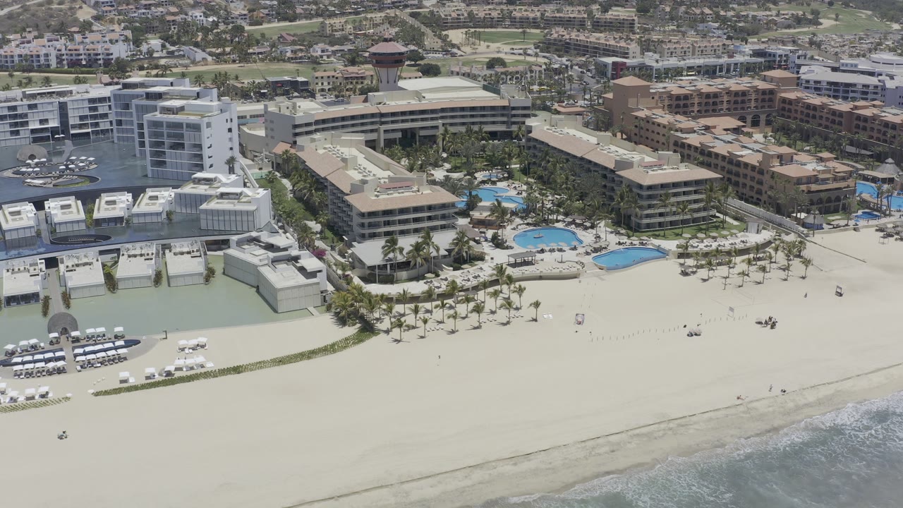 Elevated Los Cabos coastline view with the white multi-story building nearer center, blue pool shapes behind the sand, tan buildings to the right, and no visible people.