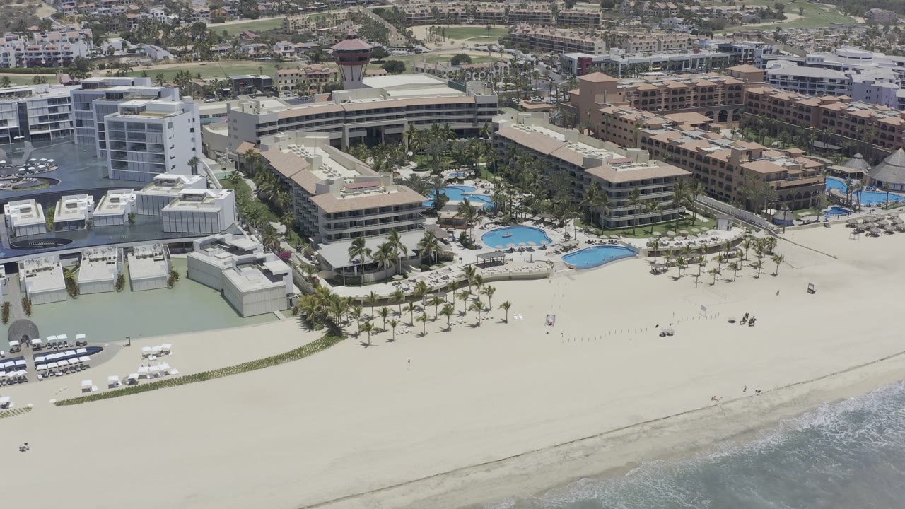 Drone-style view of coastal buildings, empty sand, palms, and pool decks.