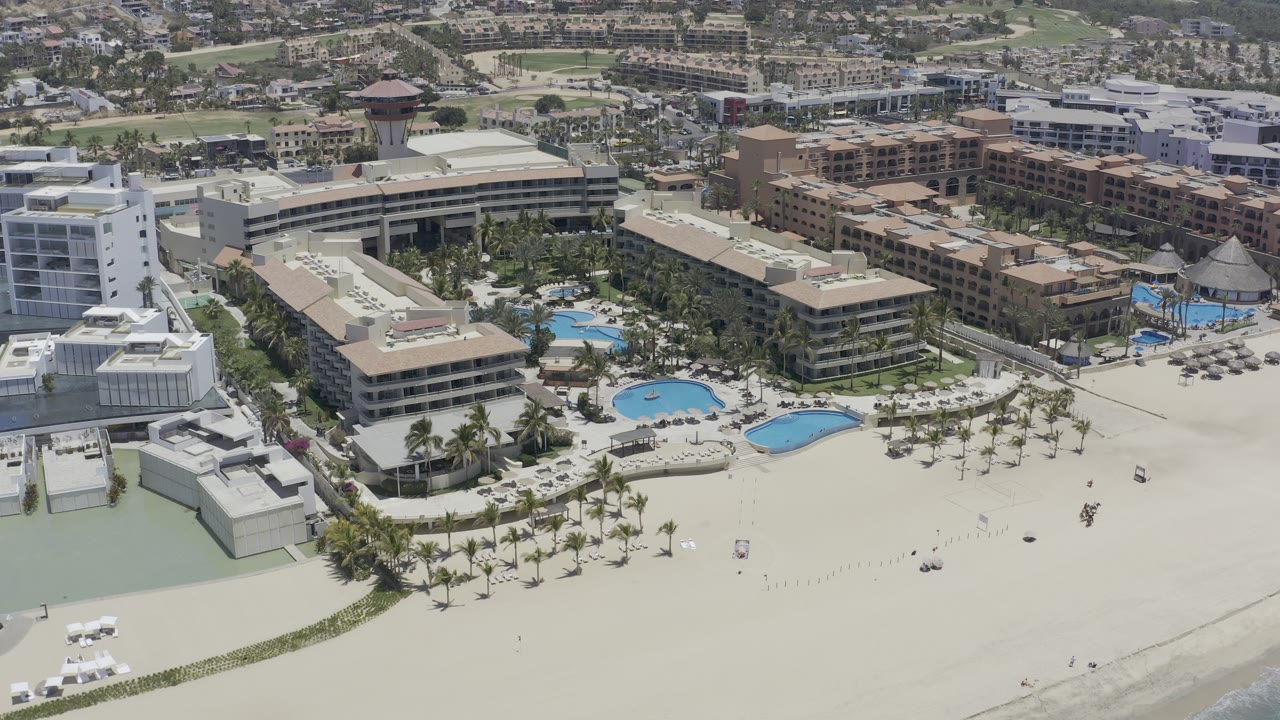Aerial view of a beachside resort with pools, palms, sand, and hotel buildings.