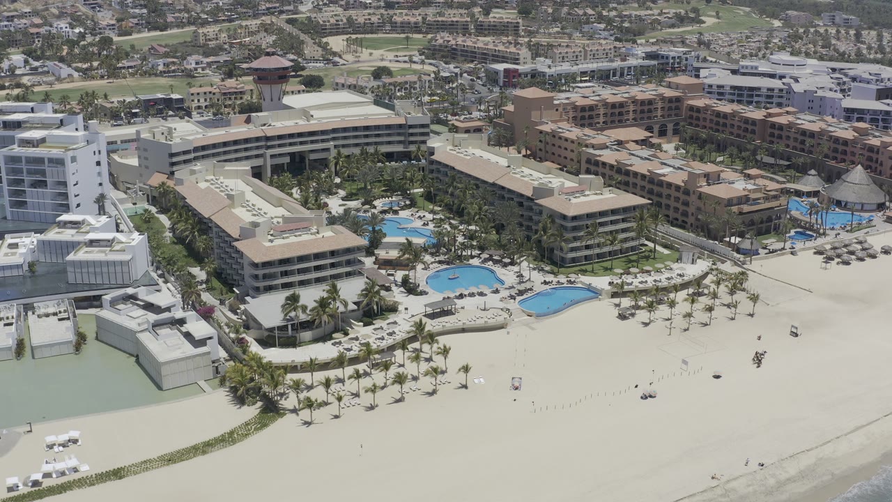 High aerial frame of a Los Cabos resort showing two curved blue pools, rows of palms on the sand, multi-story buildings, and no visible people.