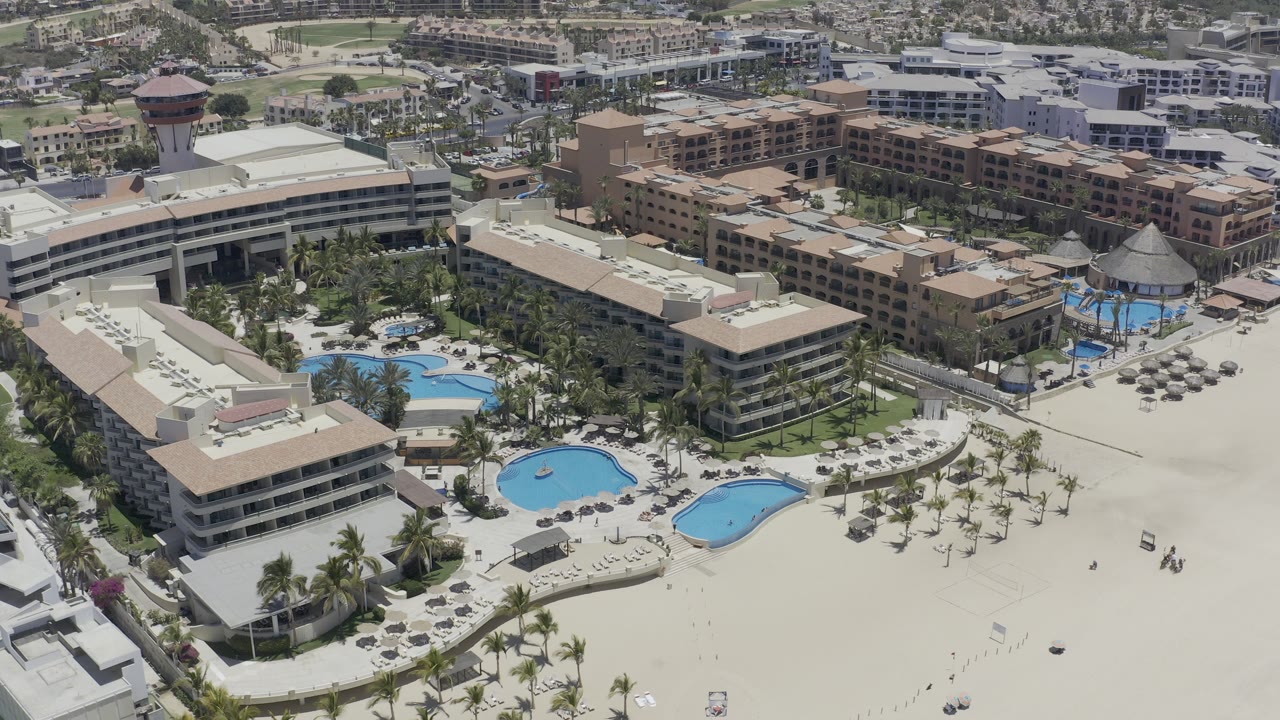 Aerial view of a beach resort with blue pools, palm trees, and tan hotel roofs.