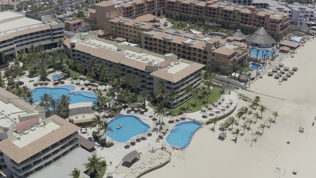 Aerial view of resort pools, palm trees, hotel buildings, and beach sand.