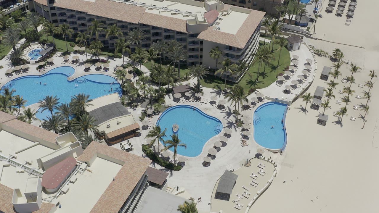 Overhead view of a Los Cabos resort pool complex with blue curved pools, palms, lounge chairs, tan-roofed buildings, and no clearly visible people.