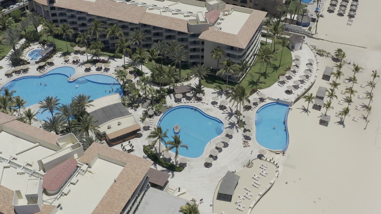 Aerial frame over the Los Cabos resort showing multiple blue pools, palm landscaping, pale walkways, rows of lounge chairs, and no clearly visible people.