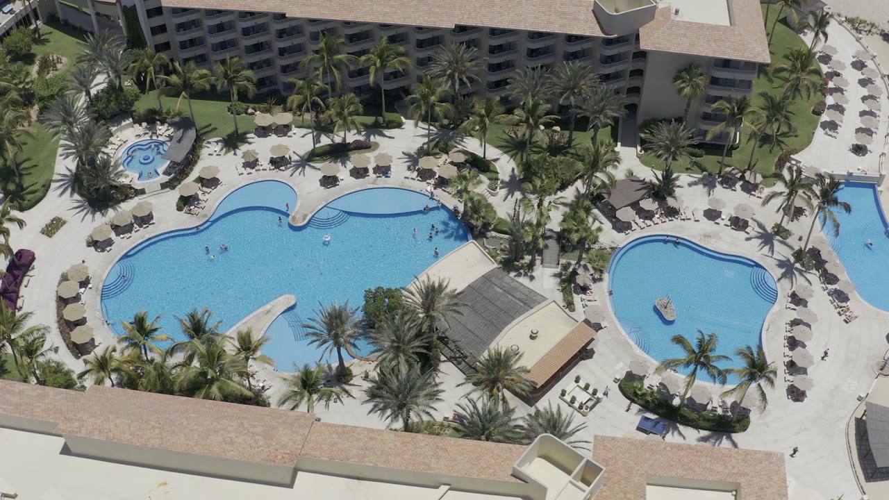 Overhead view of two resort swimming pools with palm landscaping and lounge chairs.