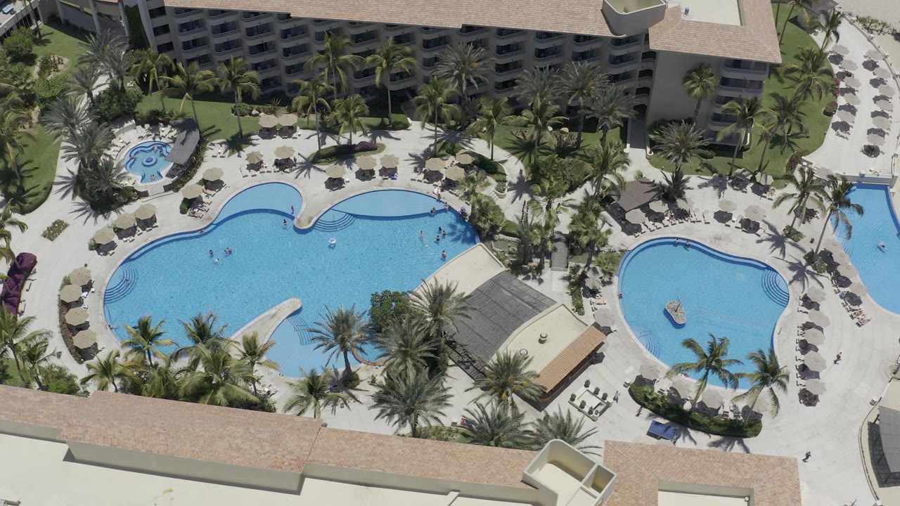 Los Cabos aerial frame with the larger pool centered, the smaller pool to the right, rows of umbrellas, palm landscaping, and hotel balconies along the top edge.