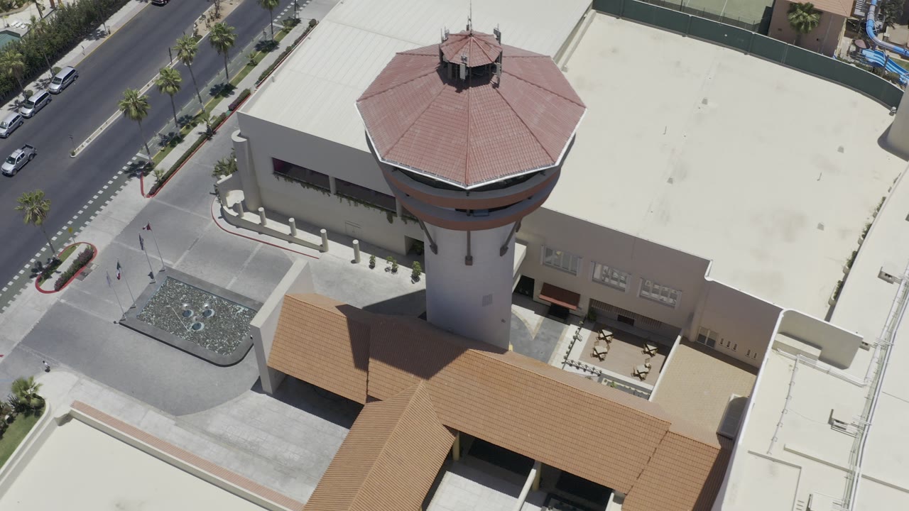 Aerial frame in Los Cabos, Mexico, looking down on a white building with a tall round tower, red roof, nearby roads, cars, palms, and small visible pedestrians.