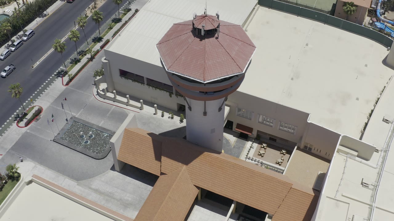 Aerial Los Cabos frame showing the red-roofed tower closer to center, with pale roof surfaces, a paved forecourt, roadside palms, cars, and tiny pedestrians below.