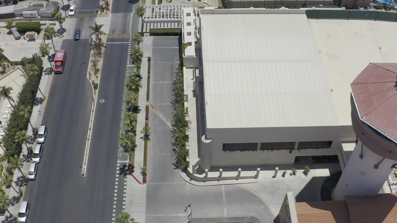 Aerial frame in Los Cabos, Mexico, looking down on the pale roof, curved tan building edge, palm-lined street, parked vehicles, and no visible people.
