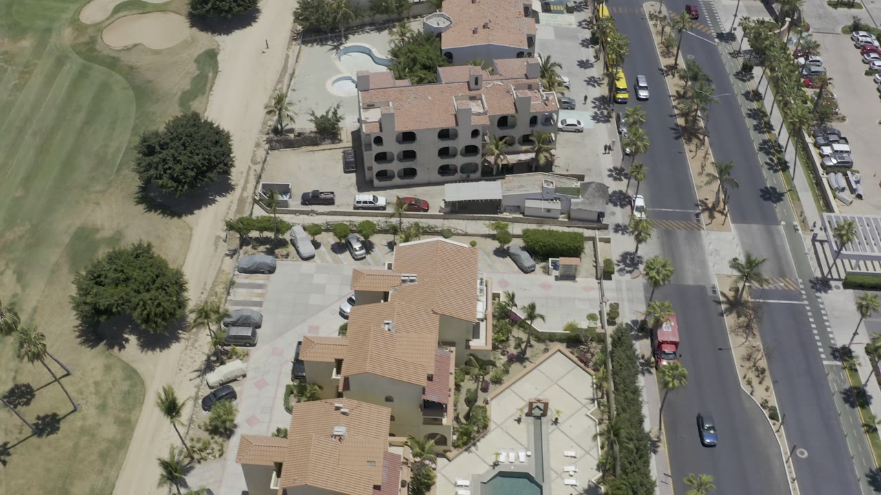 Aerial Los Cabos view with condo roofs centered between a green fairway edge, a paved road, parking areas, and no visible people.