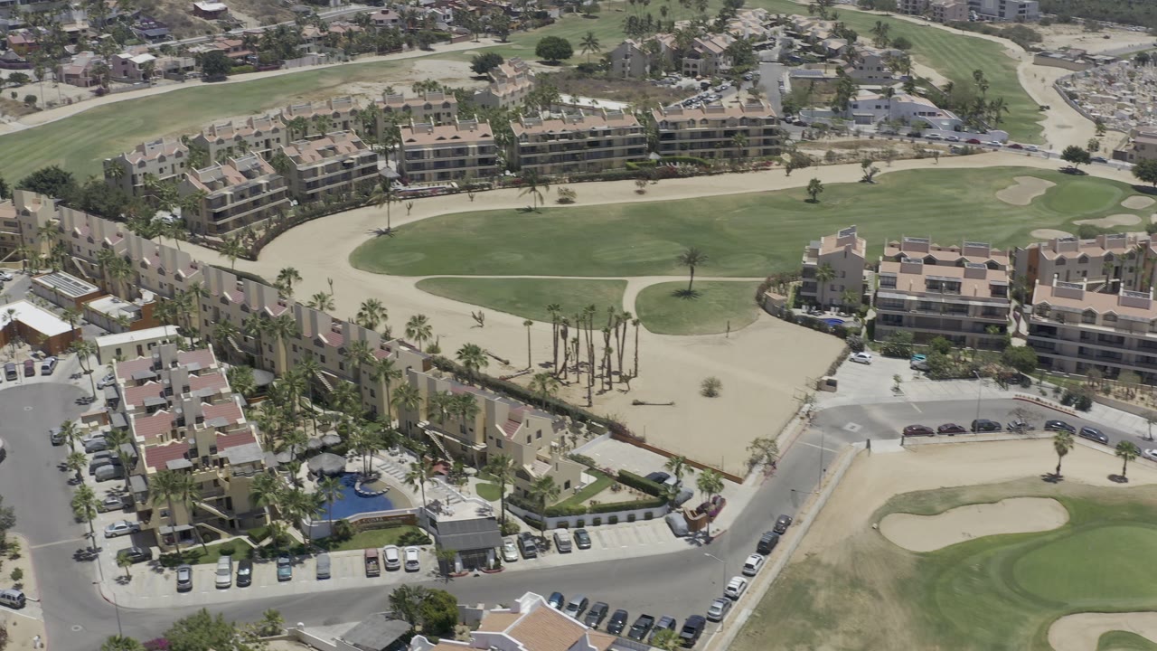 High aerial frame in Los Cabos showing manicured fairways, tan sand bunkers, multi-story resort buildings, palm-lined streets, and parking areas, with no visible people.