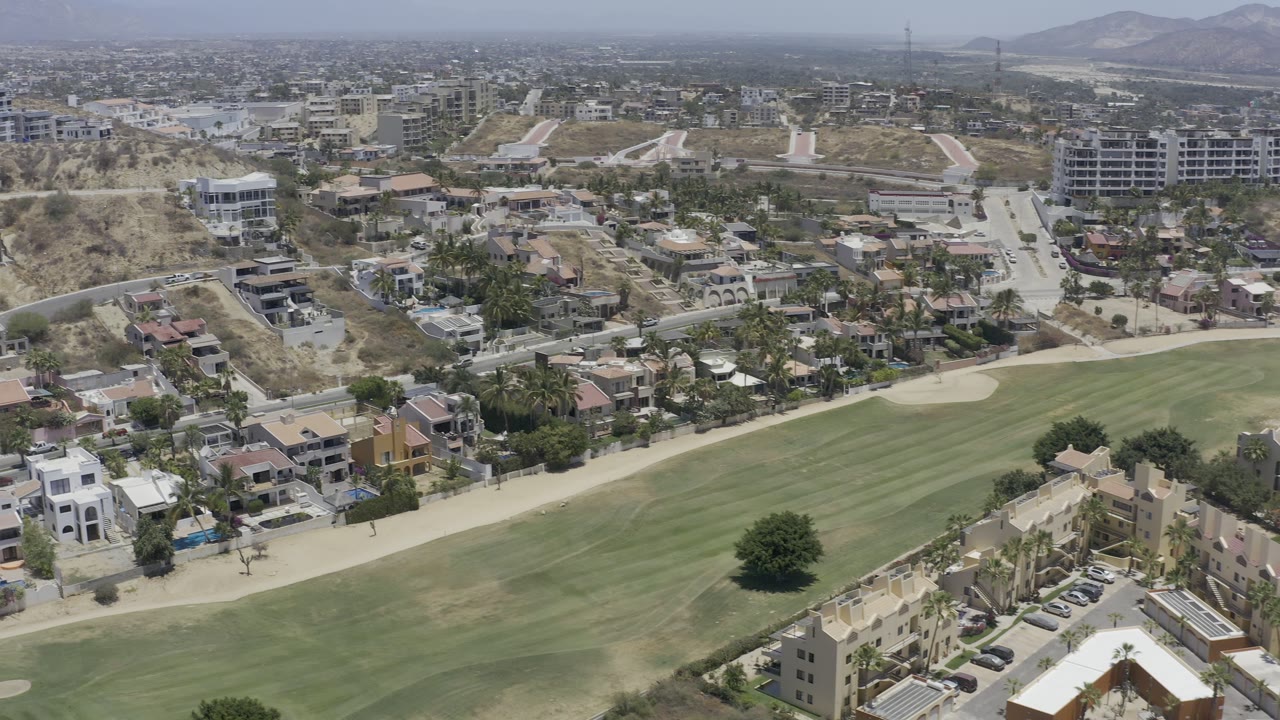 Los Cabos aerial frame with hillside residences, a long green lawn or fairway, nearby roads, scattered palm trees, small swimming pools, and no visible people.