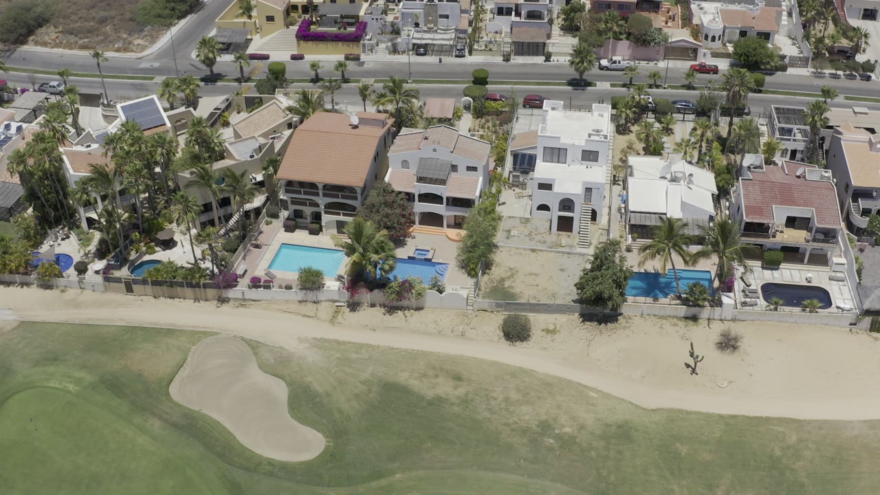 Aerial view in Los Cabos, Mexico showing beachfront condominiums, swimming pools, palm trees, sandy beach, and green turf with no visible people.