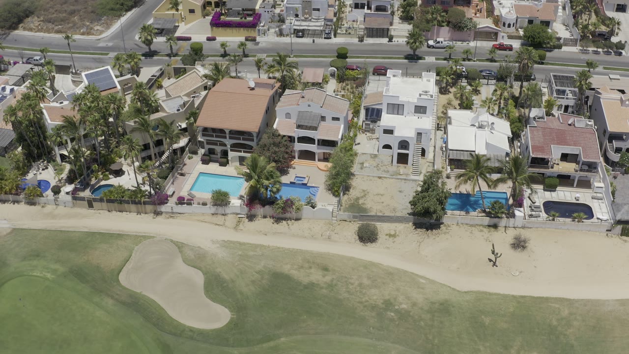 Overhead Los Cabos frame with beachfront homes, blue pools, palms, a road behind the properties, and pale sand in the lower frame with no visible people.