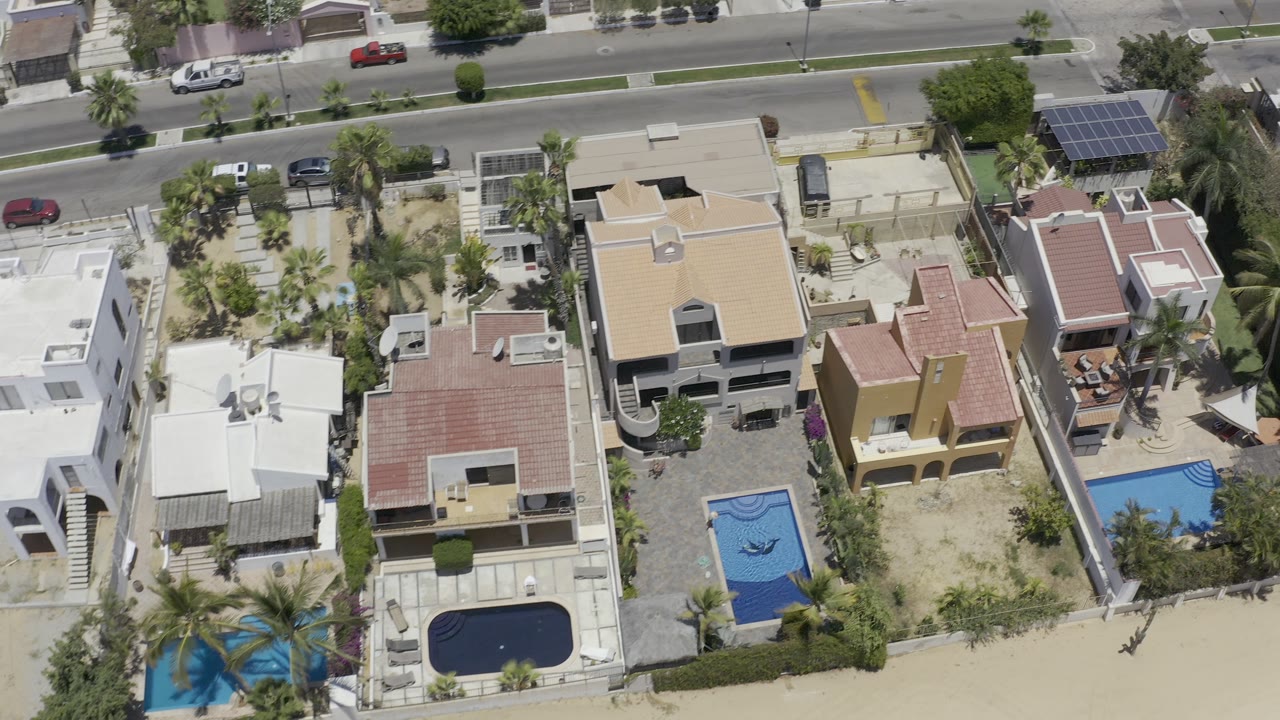 Top-down Los Cabos frame showing a row of houses, two bright blue pools, palm landscaping, a paved road with cars, and no visible people.