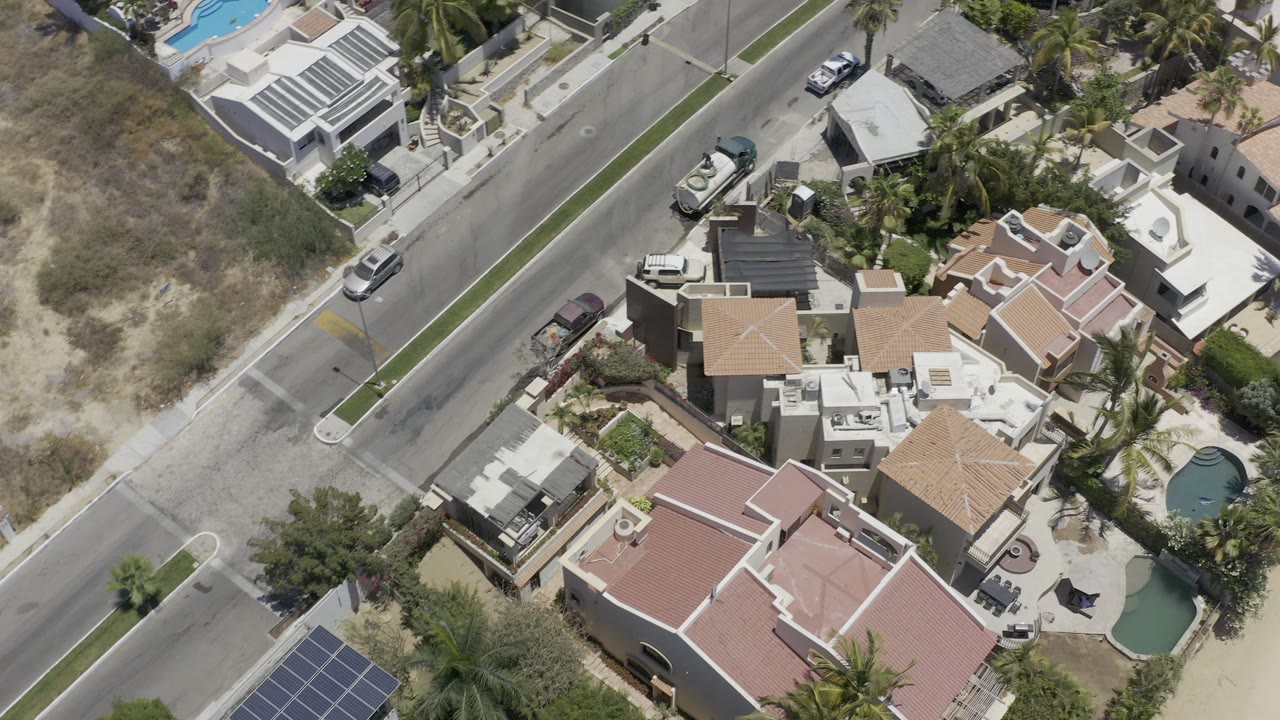 Overhead view of homes, pools, road, parked cars, palms, and dry open ground.