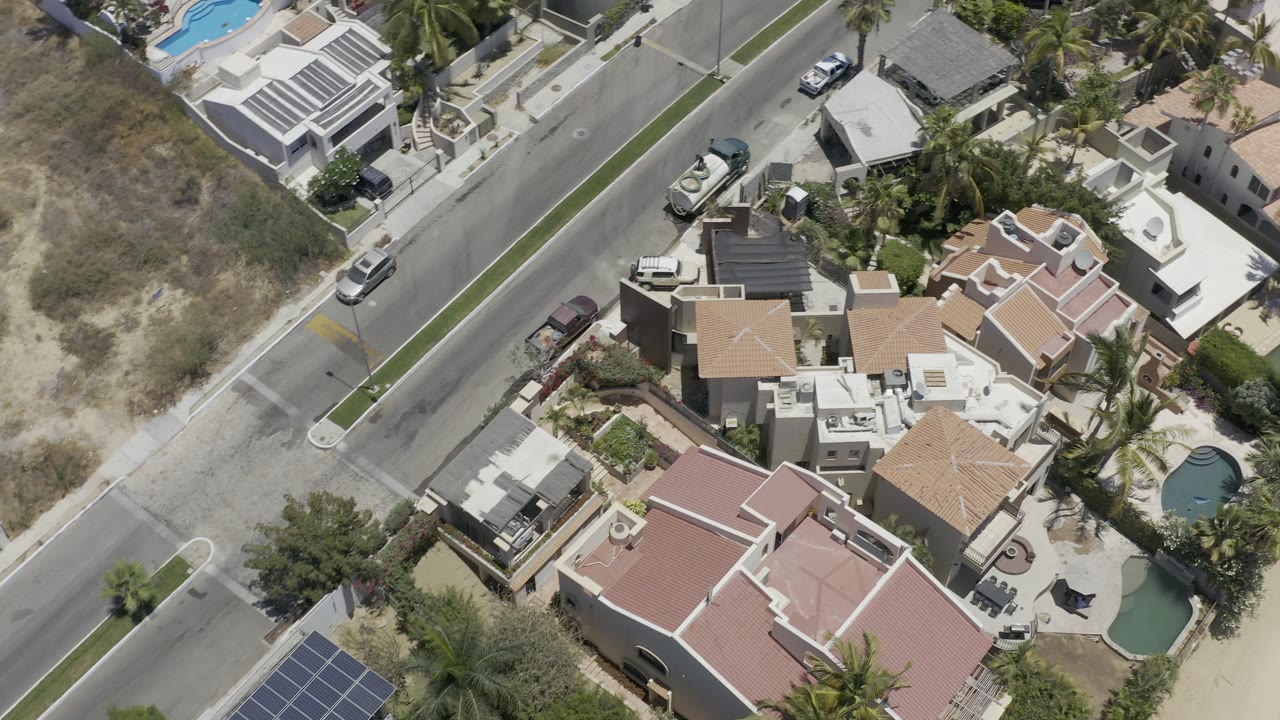 Second overhead frame in Los Cabos, Mexico with tan and white rooftops, walled yards, small pools, a paved road, parked vehicles, palms, and no visible people.