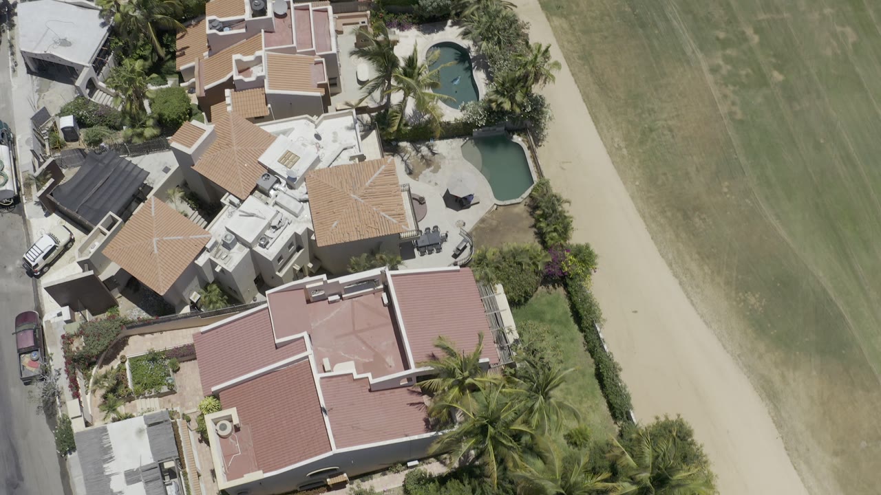 Top-down Los Cabos residential view with several pink roofed homes, walled yards, two small pools, parked cars, palms, and no visible people.