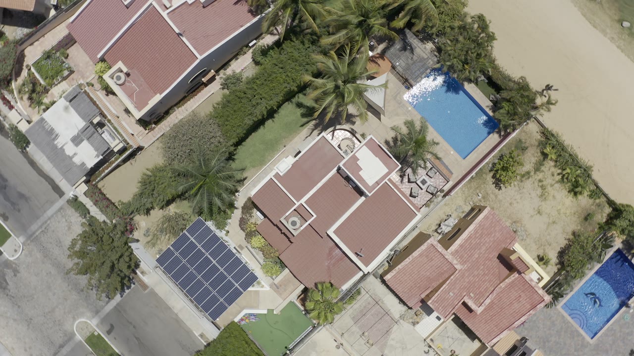 Aerial Los Cabos view with red tile roofs centered between a blue pool, palm landscaping, a road edge, and solar panels, with no visible people.