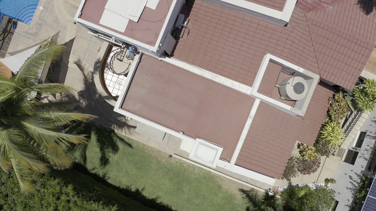 Second overhead frame in Los Cabos, Mexico keeps the red roof and white trim centered while palm fronds and their shadows cover more of the lawn, with no visible people.
