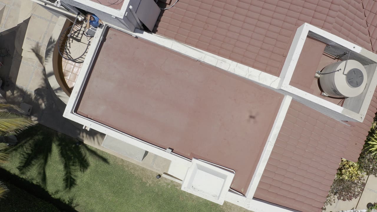 Aerial view in Los Cabos, Mexico looking down on the same red roof plane, white roof trim, nearby tile roof, palm shadow, and no visible people.