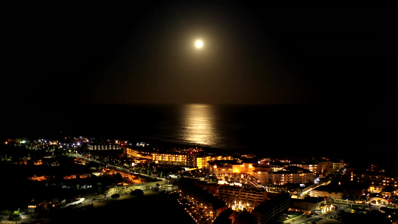 Moonlit ocean over Los Cabos, Mexico with a bright reflection on the water, illuminated waterfront buildings below, and no visible people.