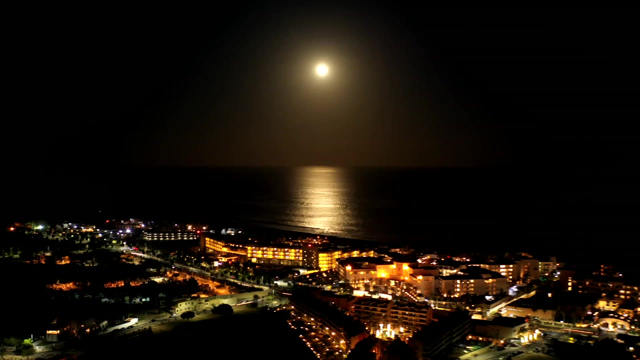 A slightly shifted night view of Los Cabos, Mexico shows the moon glowing above the ocean and warm building lights spread across the lower frame with no visible people.