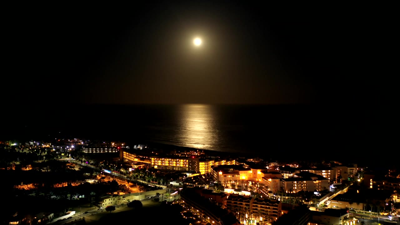 Elevated night coastline with moon over ocean and lit buildings.