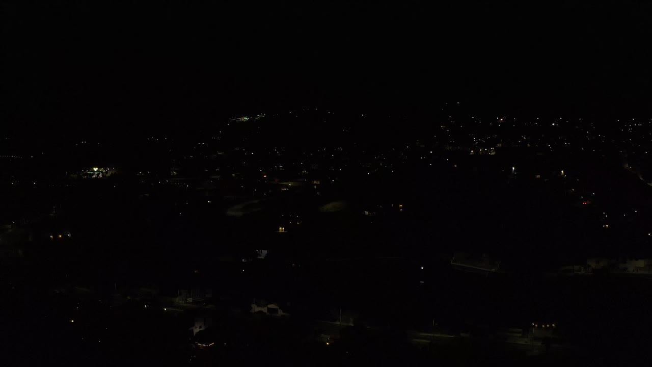 Dim high-angle Los Cabos night view with small clusters of street and building lights against deep black areas, with no visible people.