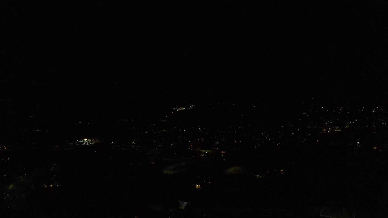 A low-exposure aerial frame of Los Cabos, Mexico, showing tiny white and warm lights spread across a dark cityscape with no visible people.
