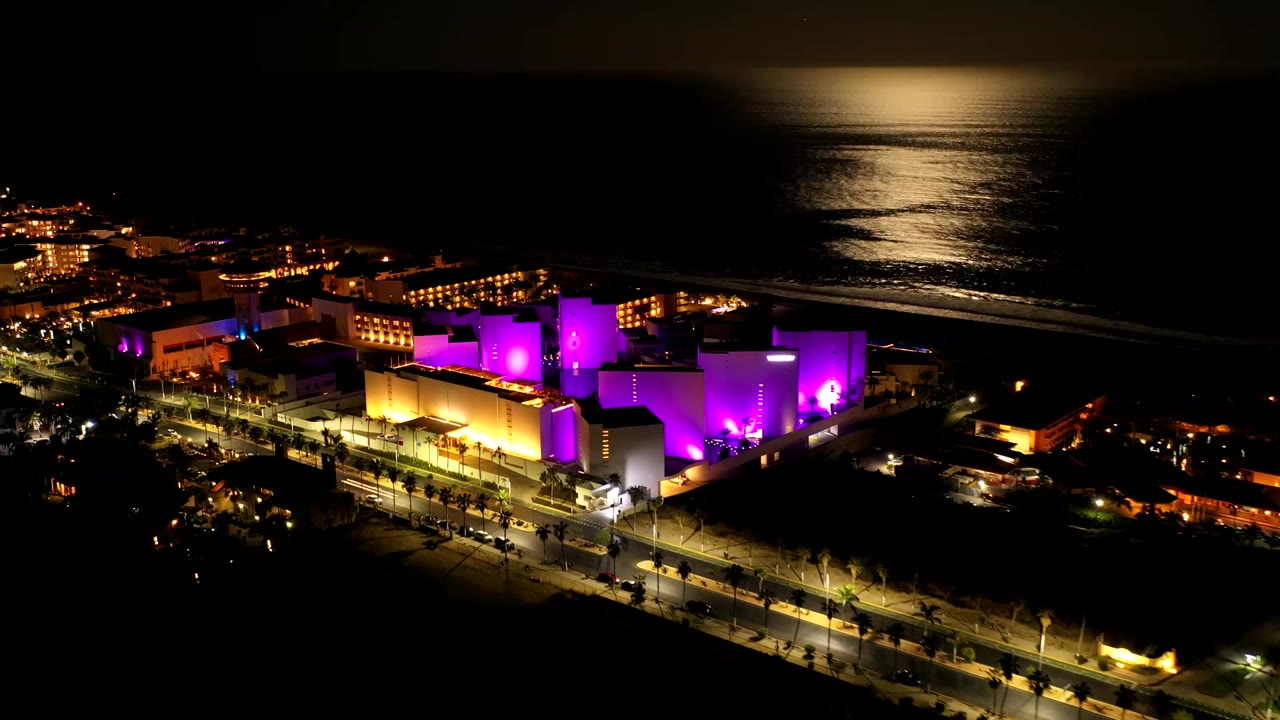 Los Cabos night aerial with the bright complex centered lower in frame, warm street lighting at left, dark shoreline beyond, and no visible people.