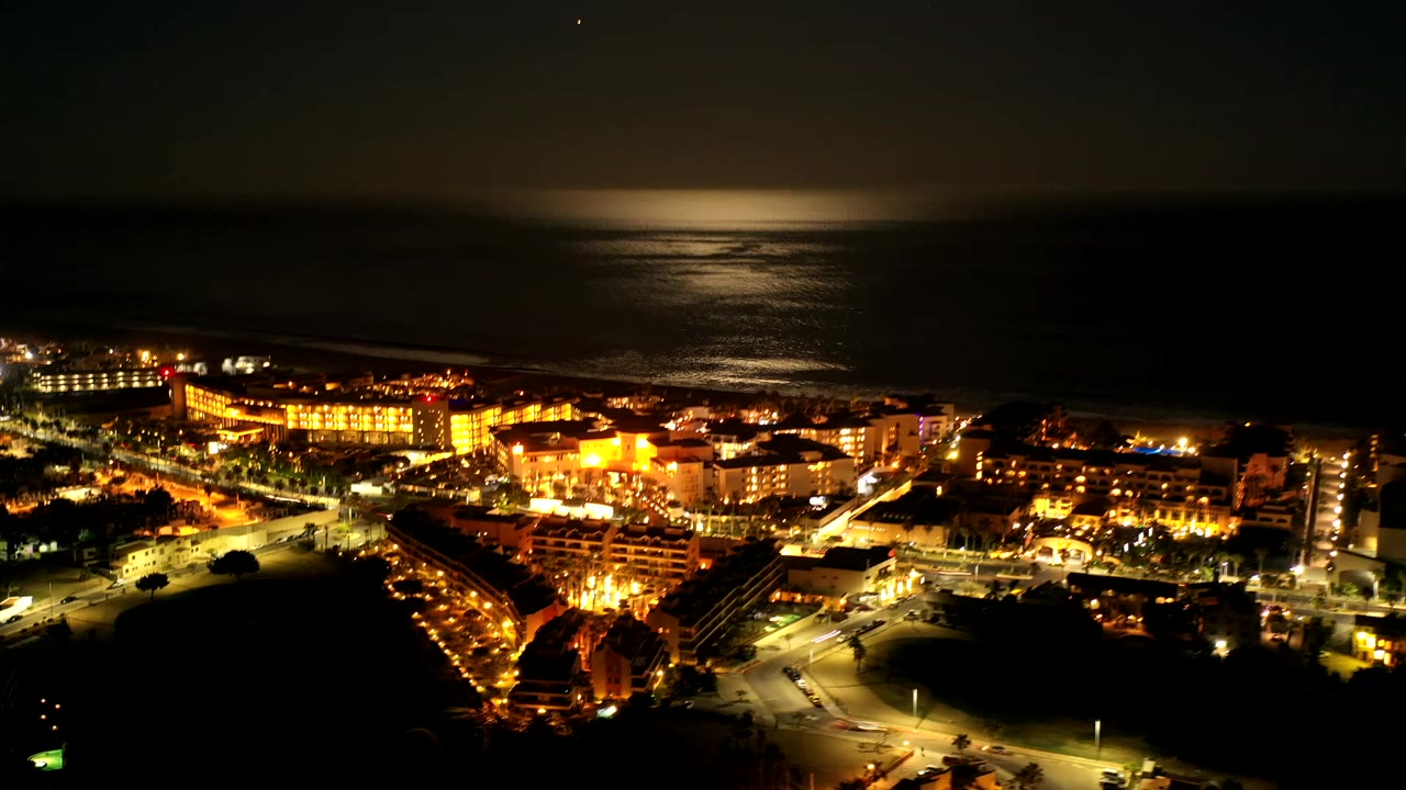 Los Cabos, Mexico night aerial with warm building lights in the lower frame, dark ocean beyond, reflected moonlight near the horizon, and no visible people.