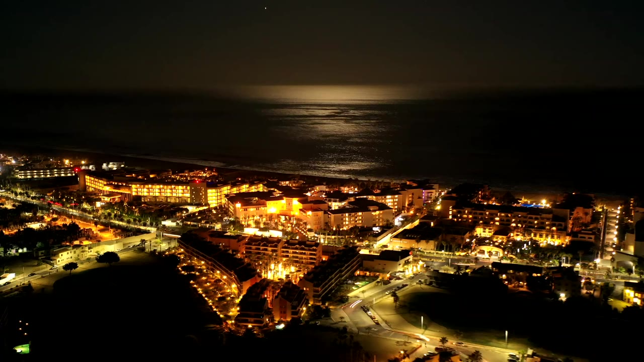 Los Cabos, Mexico appears from above at night, with bright streets and hotel-like buildings below a dark shoreline and a broad patch of moonlight on the ocean, with no visible people.