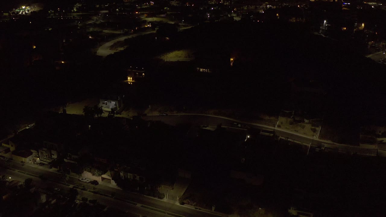 Elevated Los Cabos night frame showing a lit roadway through mostly dark residential blocks, small building lights, and no visible people.