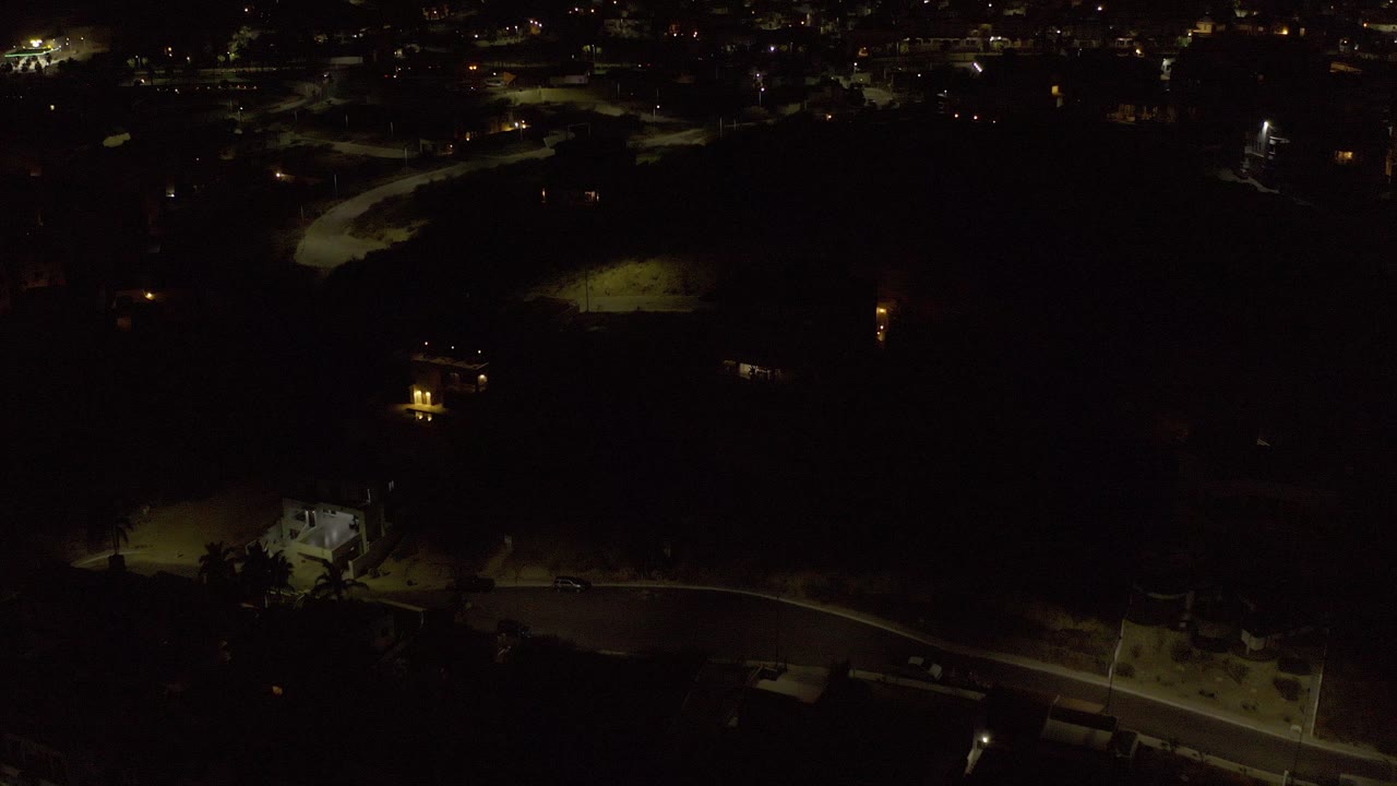 High night view over Los Cabos, Mexico, with dark roads, small pools of streetlight, distant city lights, and no visible people.