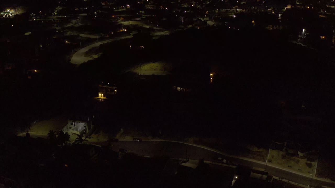 Los Cabos night aerial with a curved road lit by yellow streetlights, scattered homes, dark tree cover, and no visible people.