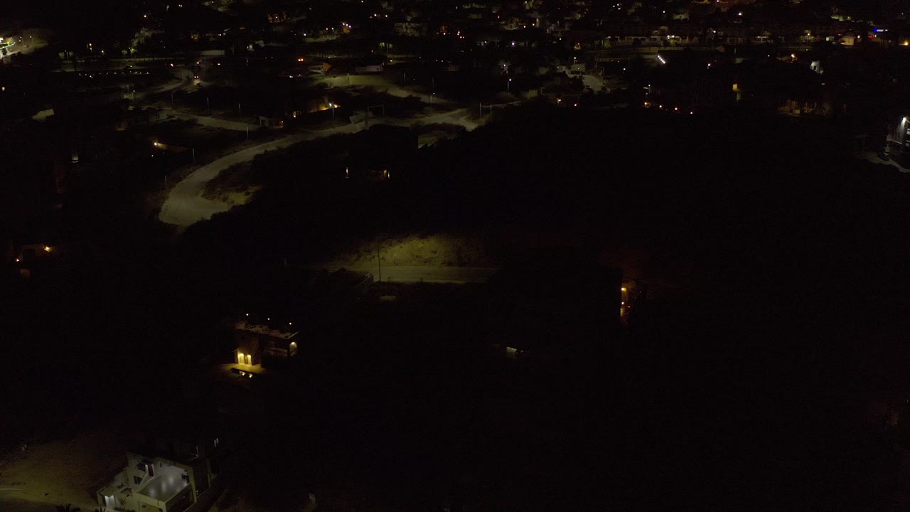 Dark aerial view over Los Cabos, Mexico, with scattered lights across the upper background, olive green-lit ground patches, a bright structure near the lower edge, and no visible people.