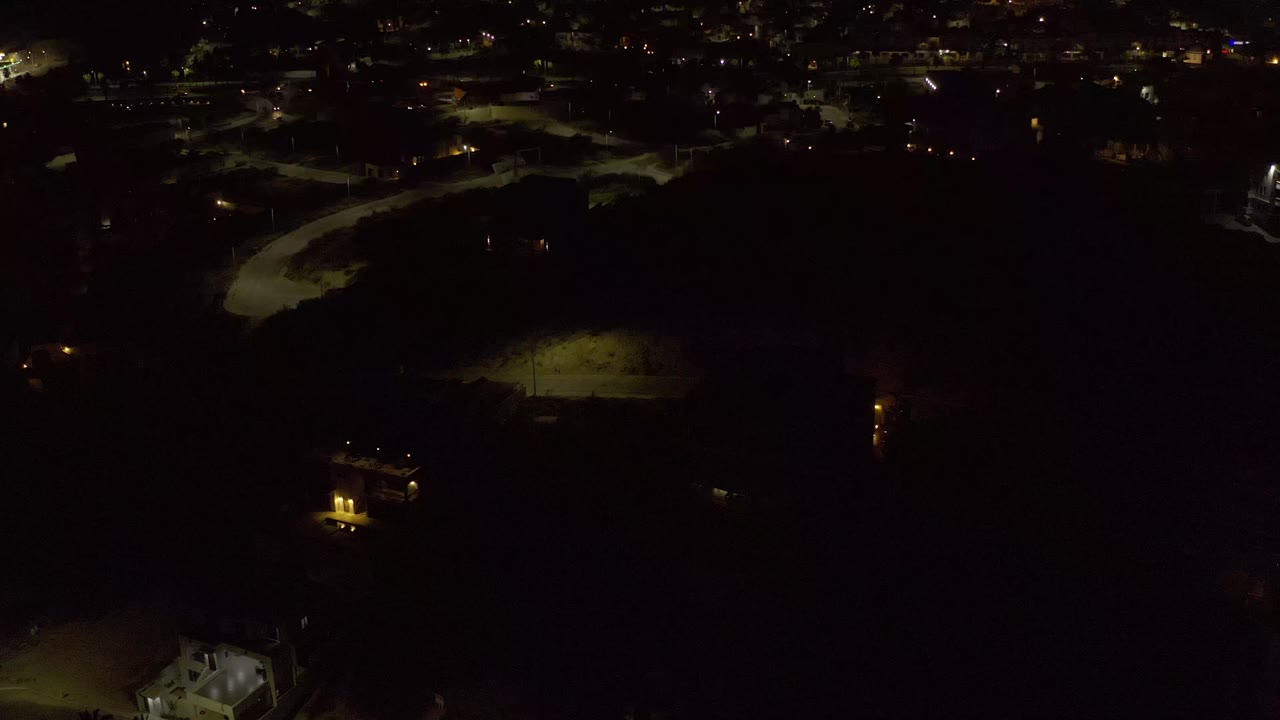 Aerial night frame from Los Cabos, Mexico, showing a band of distant city lights, a greenish lit area near center left, dark open foreground, and no visible people.